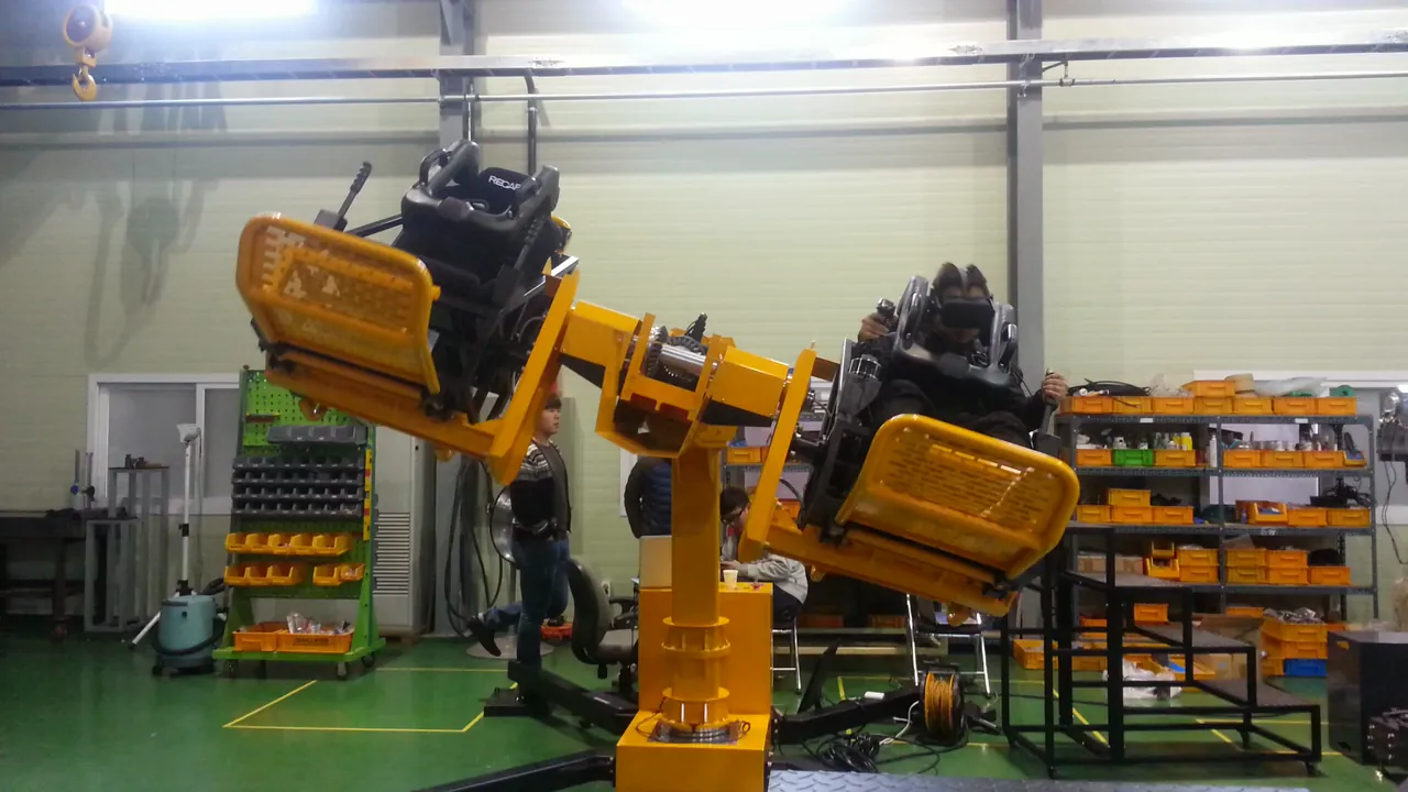 A yellow robot arm with two seats tilting with riders on board inside a factory